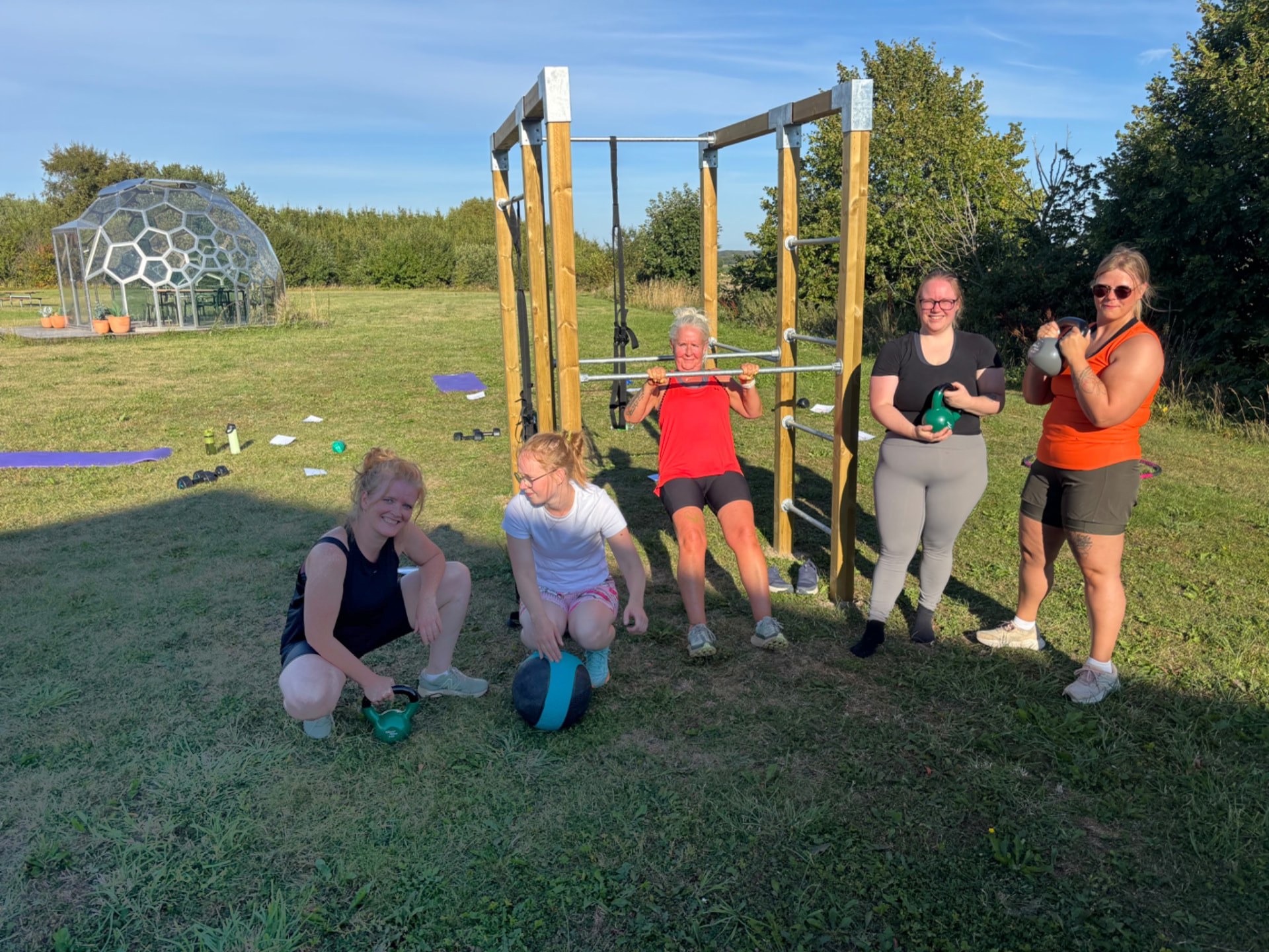 Fem deltagere fra holdtræningen samlet ved det udendørs træningsstativ med kettlebells og medicinbold